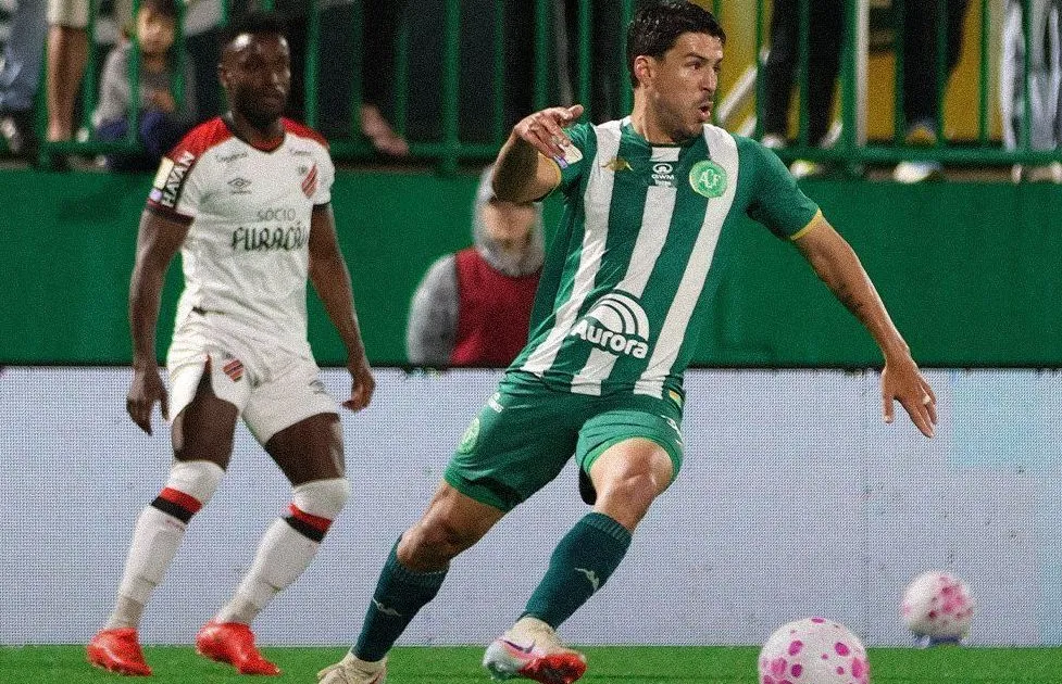 Jogador de camisa verde e listras brancas conduz a bola em campo, enquanto um adversário de branco e vermelho observa ao fundo