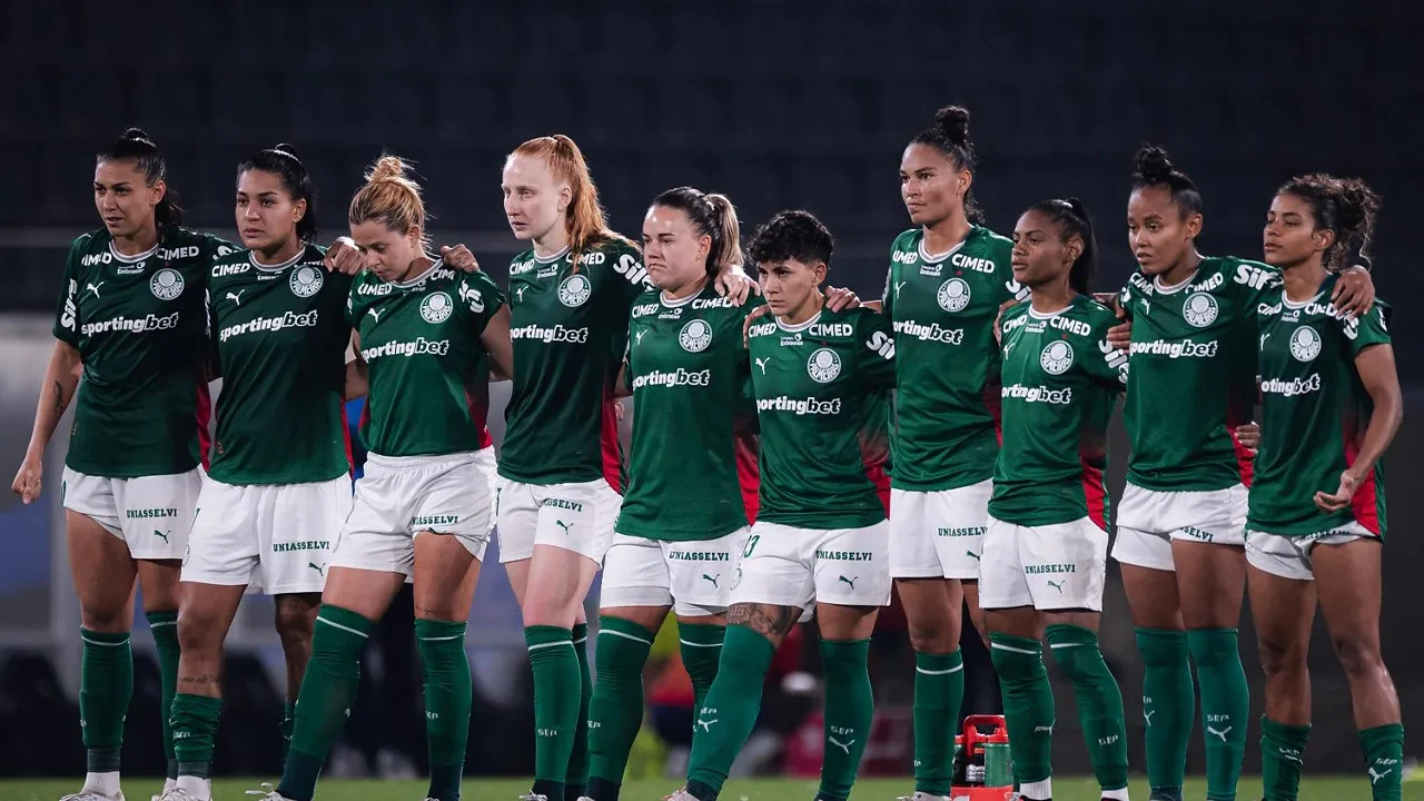 Jogadoras do Palmeiras, de uniforme verde e branco, enfileiradas lado a lado com os braços dados em um estádio à noite