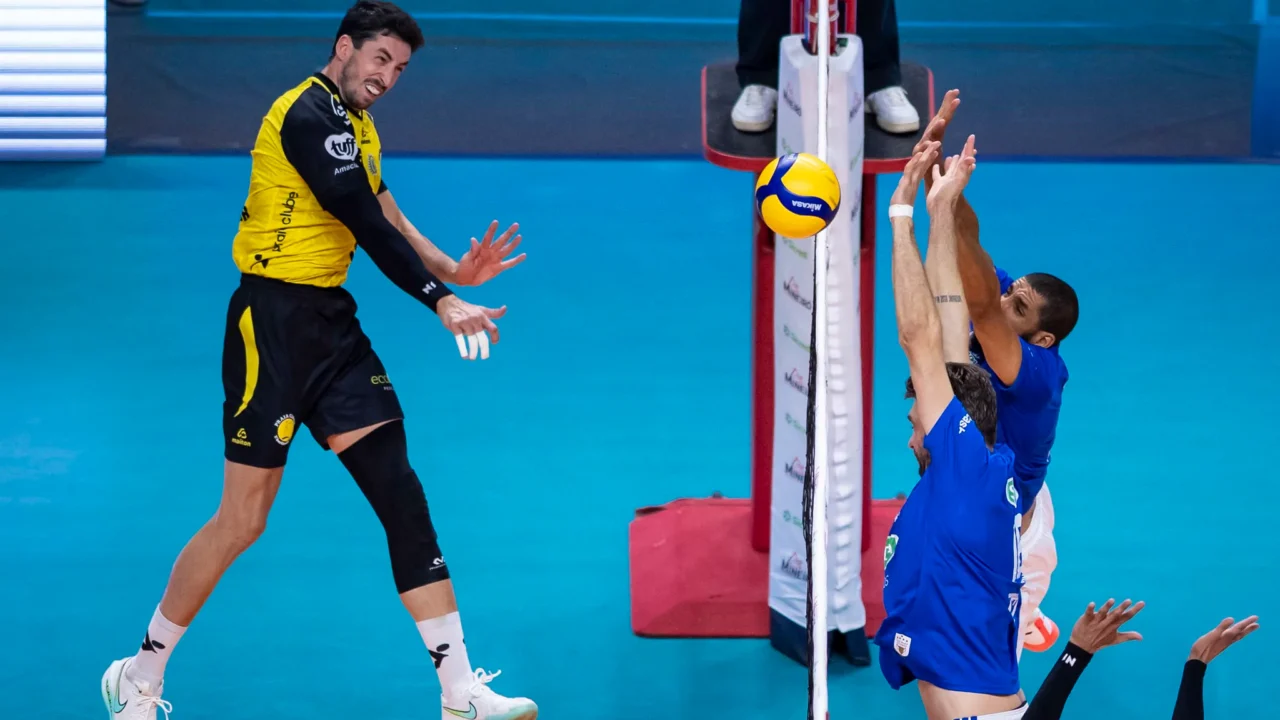Jogador de camisa amarela e preta ataca a bola sobre a rede, enquanto dois adversários de azul tentam o bloqueio em partida de vôlei