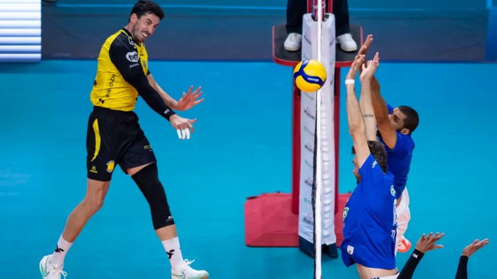 Jogador de camisa amarela e preta ataca a bola sobre a rede, enquanto dois adversários de azul tentam o bloqueio em partida de vôlei
