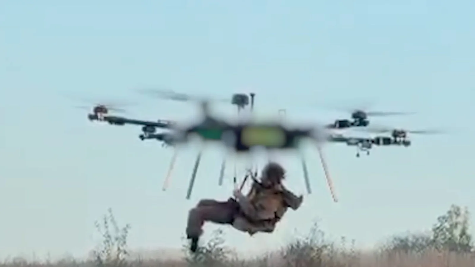 Drone pesado desce perto do chão carregando o soldado preso a um suporte, com vegetação ao fundo