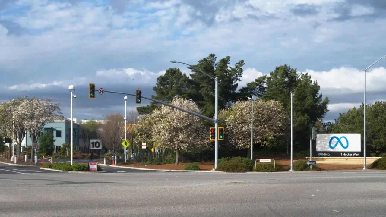 Entrada da sede da Meta em Menlo Park, na Califórnia, com a placa do logo da empresa e o endereço 1 Hacker Way, vista do outro lado de um cruzamento com semáforos