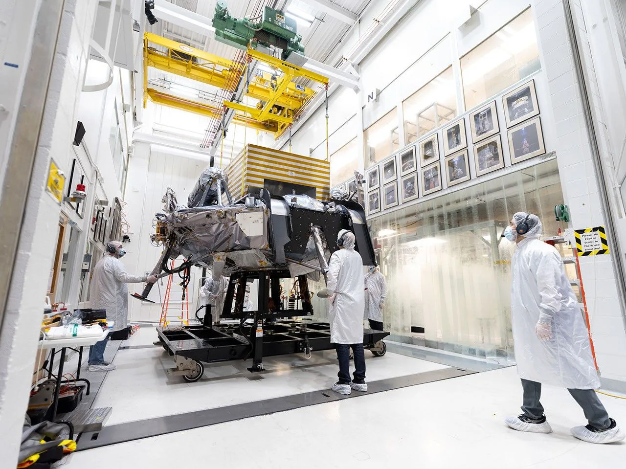 Na Figura A, engenheiros e técnicos movem a Griffin-1 por um corredor que leva ao Simulador Espacial de 25 Pés, em 31 de agosto. O corredor é ladeado por fotografias de algumas das espaçonaves históricas que foram testadas na instalação desde os anos 1960.