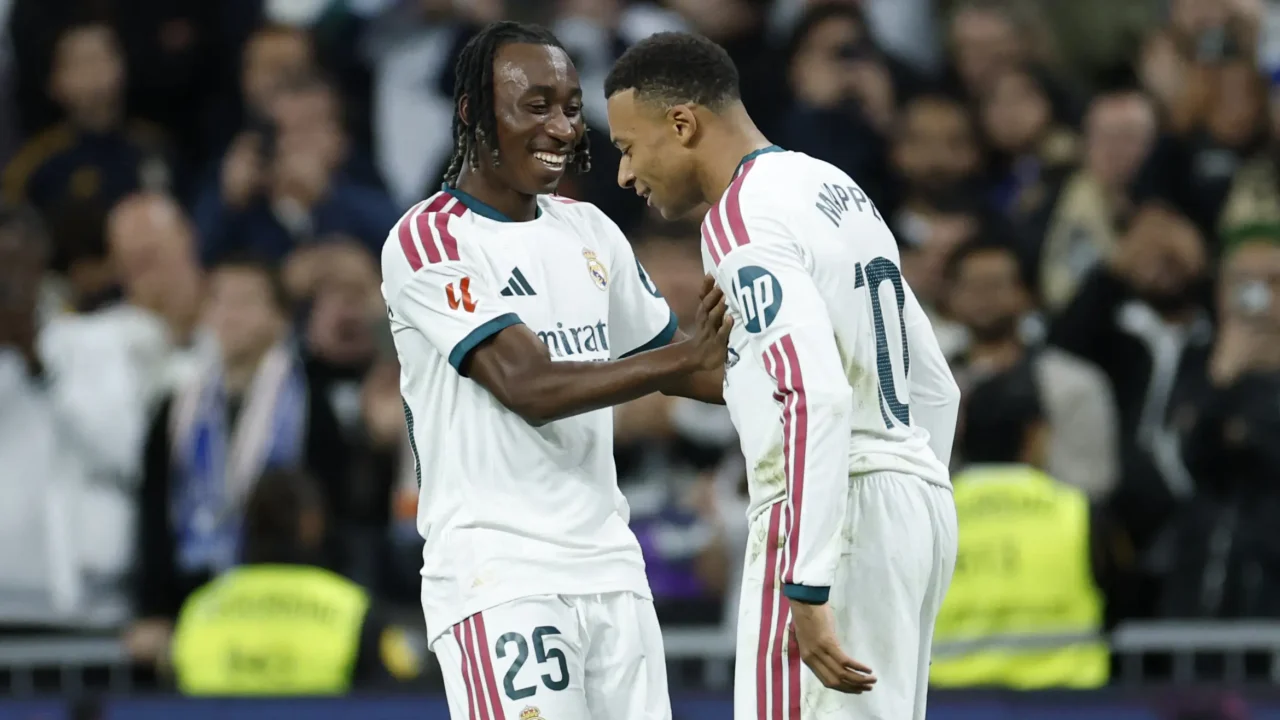Jogadores do Real Madrid comemoram com sorriso e abraço em campo, com torcida desfocada ao fundo; o da direita tem "MBAPPÉ" e número 10 nas costas
