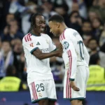 Jogadores do Real Madrid comemoram com sorriso e abraço em campo, com torcida desfocada ao fundo; o da direita tem "MBAPPÉ" e número 10 nas costas