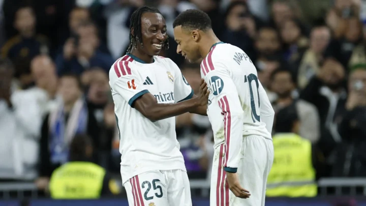 Jogadores do Real Madrid comemoram com sorriso e abraço em campo, com torcida desfocada ao fundo; o da direita tem "MBAPPÉ" e número 10 nas costas