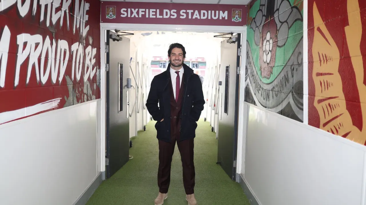 Homem de terno e gravata em corredor de estádio com gramado sintético, posicionado diante de entrada com placa identificando o local