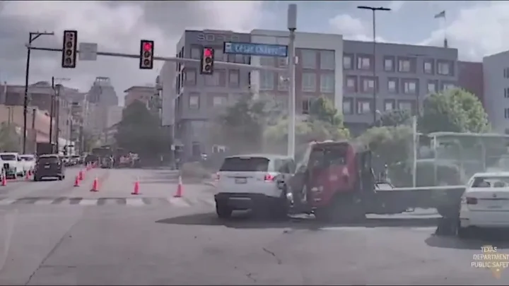 Câmera da viatura do DPS mostra o cruzamento da batida: uma SUV branca e um caminhão-plataforma vermelho colados, poeira no ar, sinais vermelhos e a placa da César Chávez no centro de San Antonio