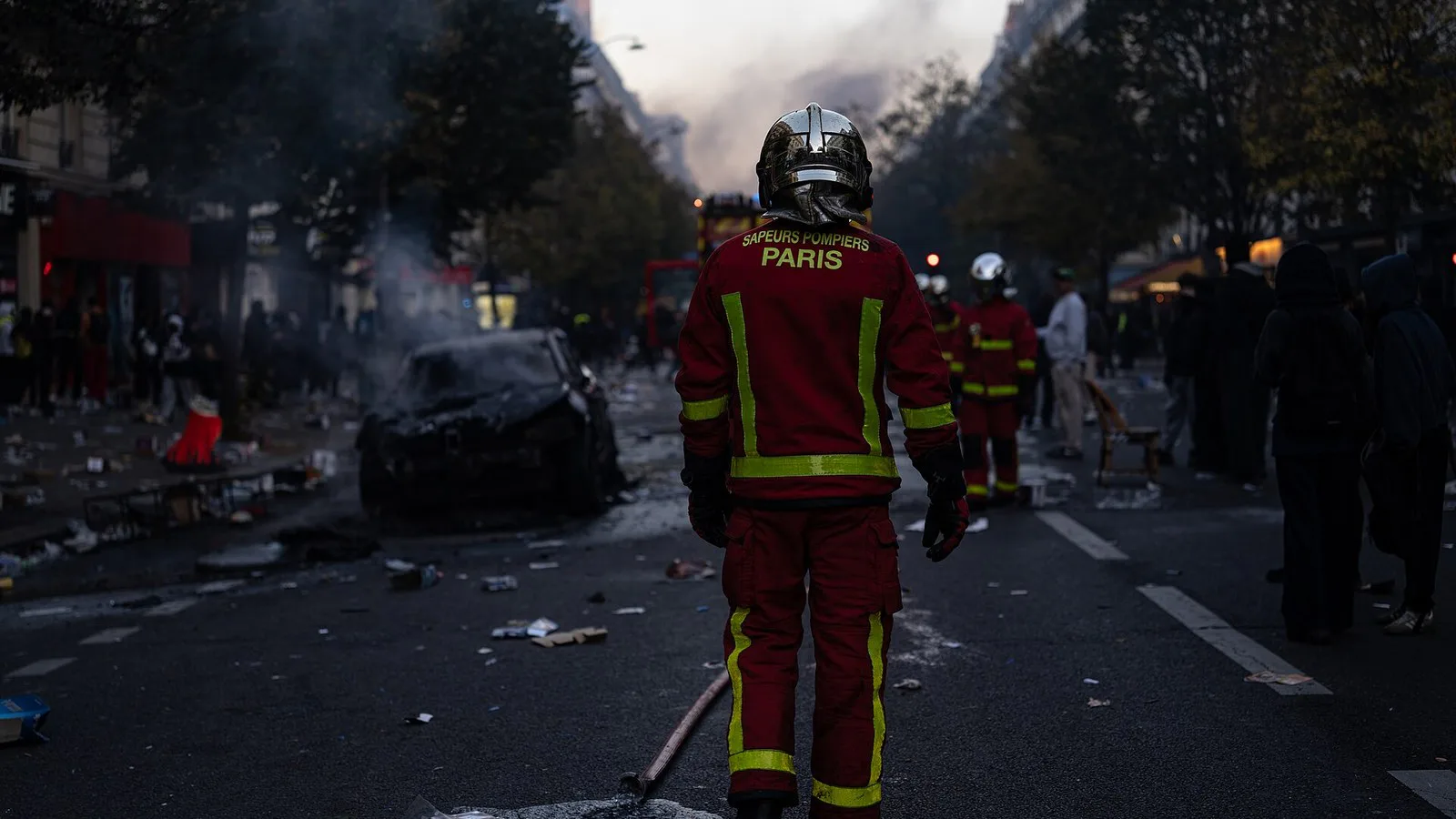 Bombeiro de Paris caminha por uma rua com destroços e um carro queimado, entre grupos de pessoas, em 17 de outubro de 2026