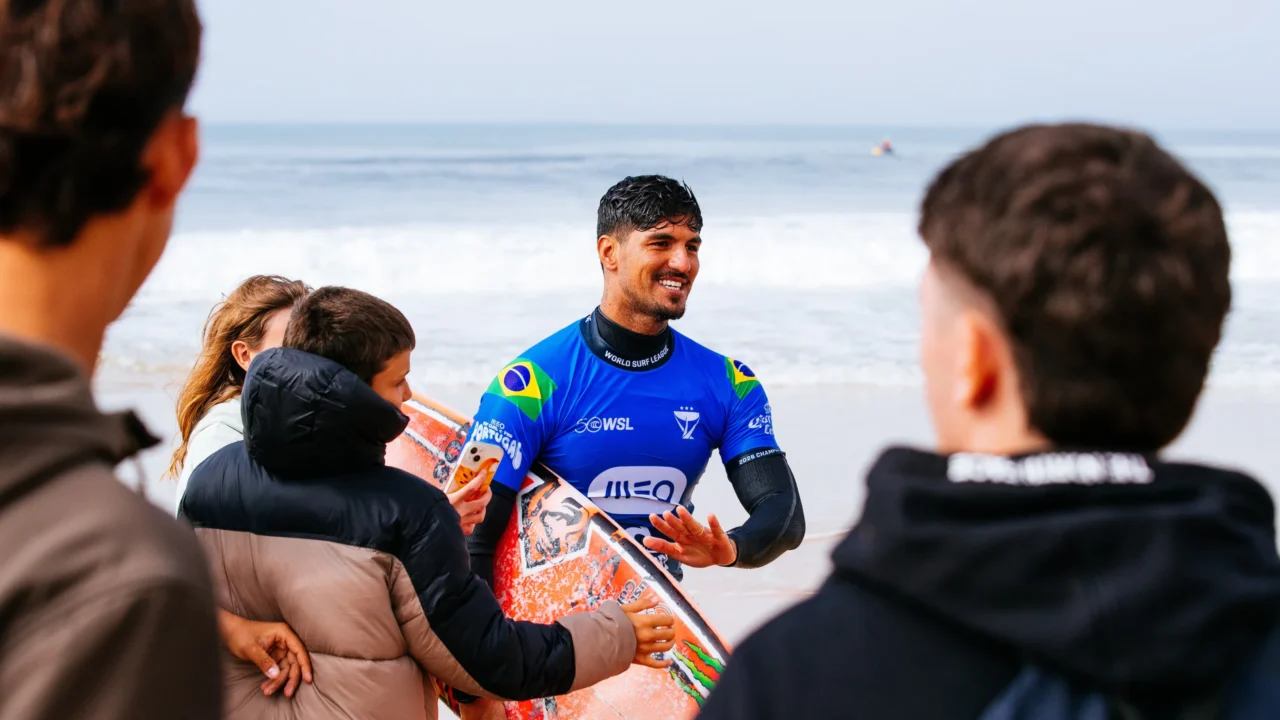 Surfista de camiseta azul com bandeira do Brasil conversa sorridente com jovens na praia, com prancha laranja nas mãos