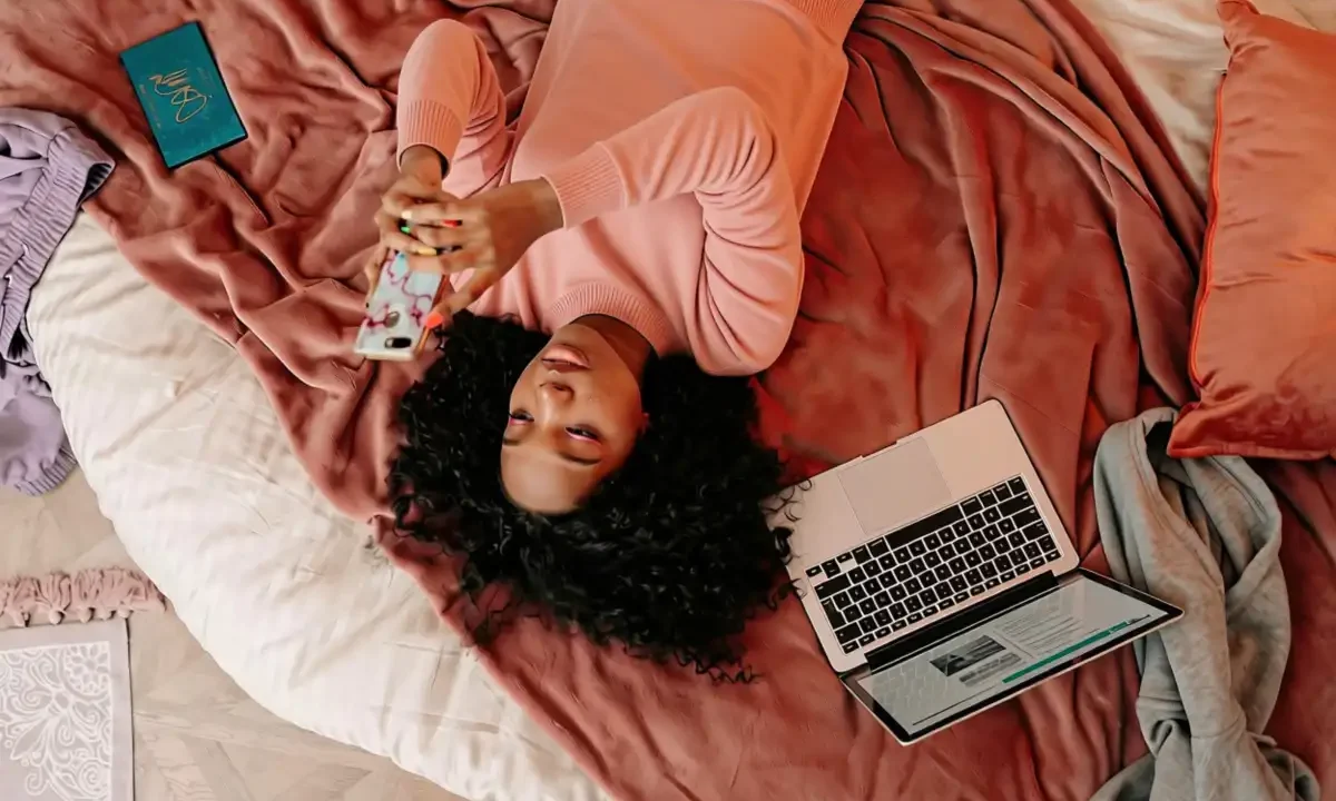 Mulher deitada em cama com roupas rosa segura smartphone enquanto usa laptop, cercada por livro, caderno e outros itens de estudo