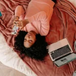 Mulher deitada em cama com roupas rosa segura smartphone enquanto usa laptop, cercada por livro, caderno e outros itens de estudo