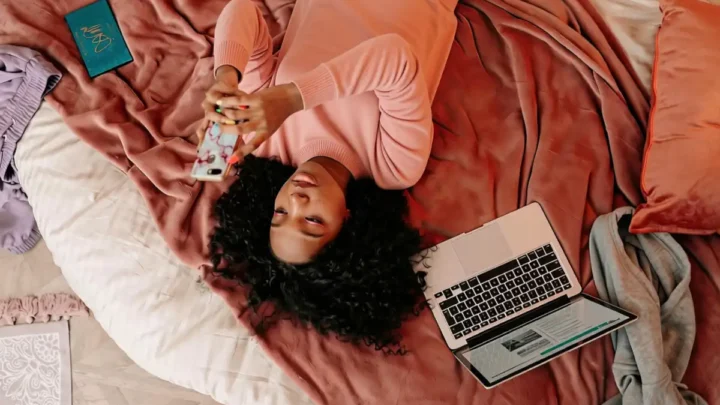 Mulher deitada em cama com roupas rosa segura smartphone enquanto usa laptop, cercada por livro, caderno e outros itens de estudo