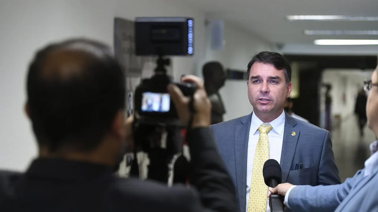 Flávio Bolsonaro, de terno cinza e gravata amarela, fala a repórteres e câmeras em um corredor, em foto de maio de 2022