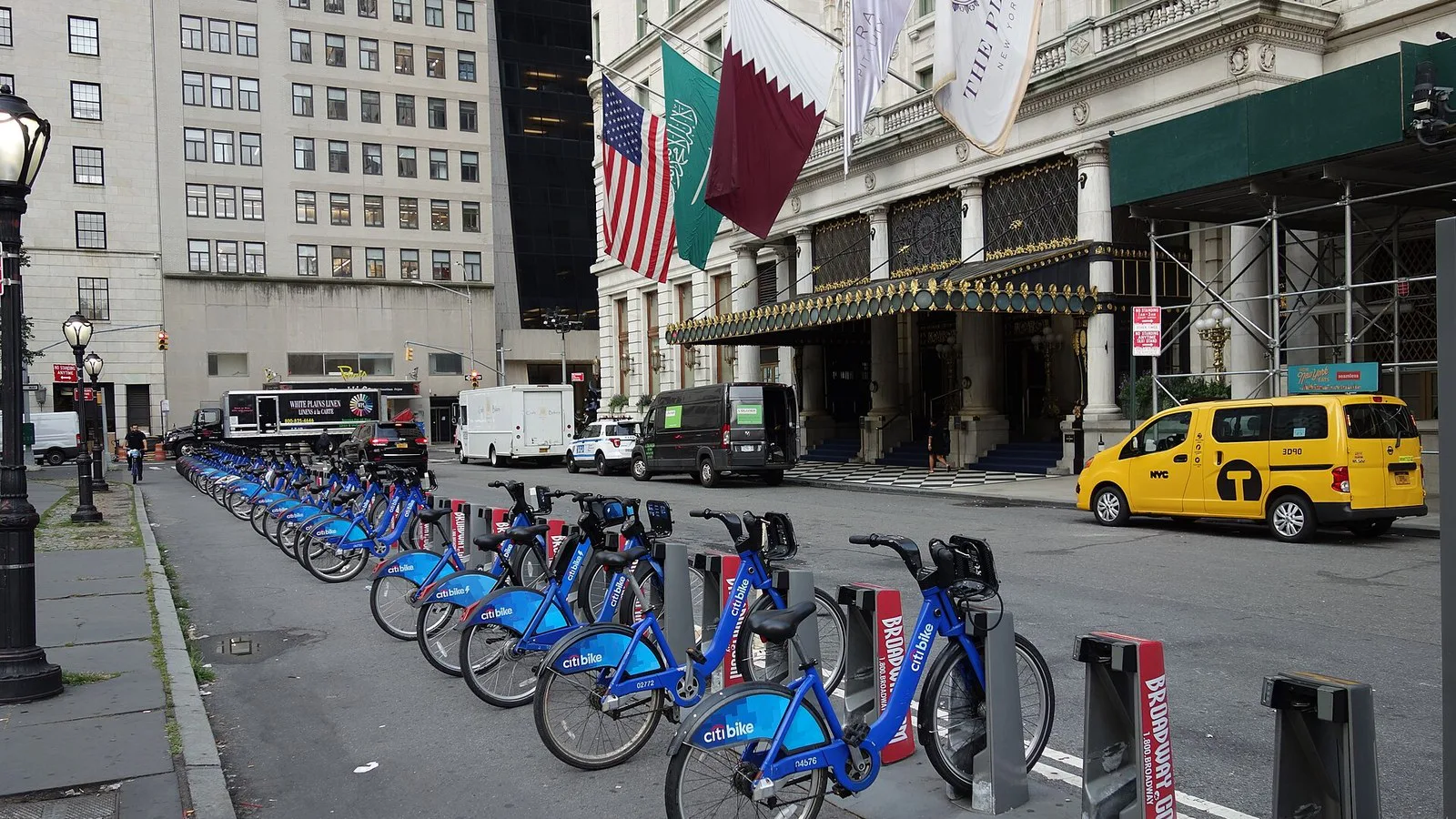 Bicicletas azuis de aluguel enfileiradas na Grand Army Plaza, com a entrada do Hotel Plaza, as bandeiras e um táxi amarelo ao fundo