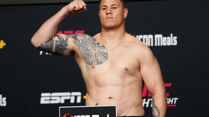 Homem sem camisa, com tatuagens no braço e no peito, flexiona o bíceps em frente a painel com logos da ESPN e do UFC, sobre placa da Iron Meals no pódio de pesagem