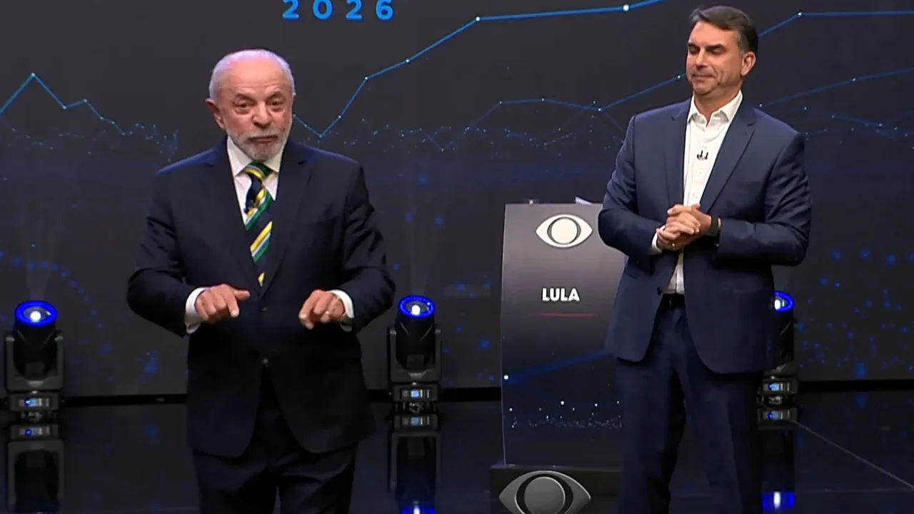 Flávio Bolsonaro e Lula em pé no palco do debate da Band, diante do painel com linhas azuis