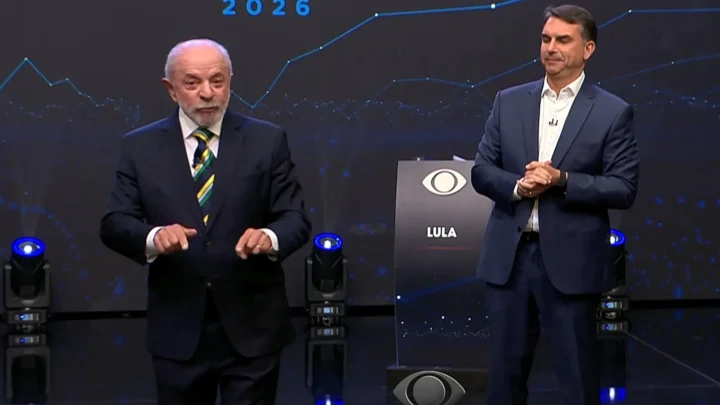 Flávio Bolsonaro e Lula em pé no palco do debate da Band, diante do painel com linhas azuis