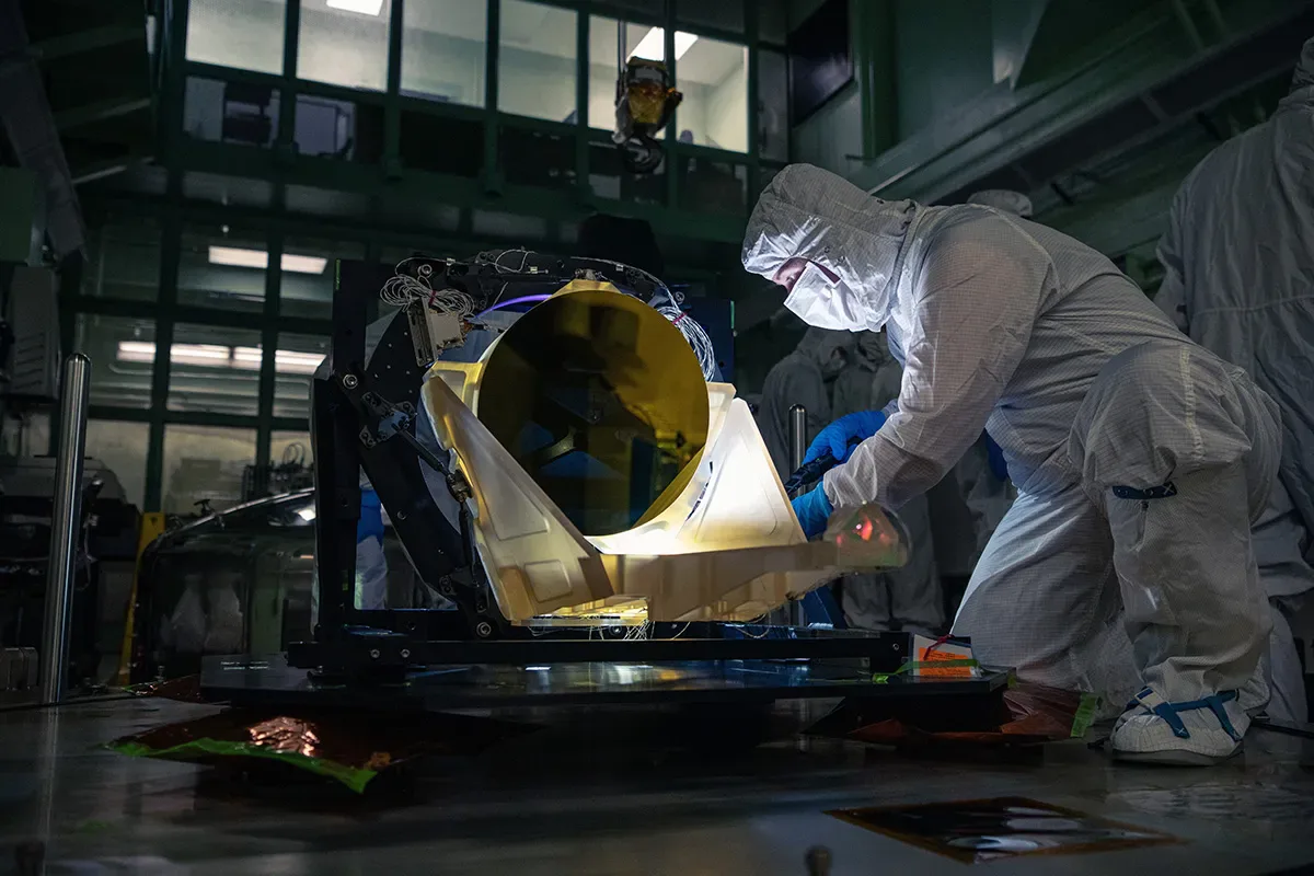 Um técnico em uma sala limpa no NASA Goddard inspeciona o telescópio protótipo do LISA entregue pela L3Harris em maio de 2024. Todo o telescópio é feito de um vidro-cerâmico de cor âmbar que resiste a mudanças de forma em uma ampla faixa de temperatura. A superfície do espelho é revestida com ouro.