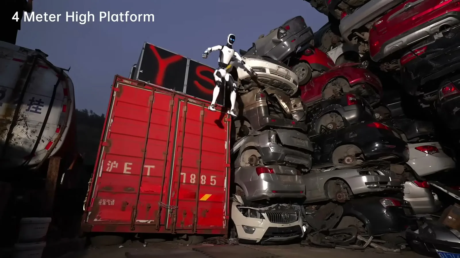 O robô H2-X no alto de um contêiner vermelho, ao lado de uma pilha de carros num ferro-velho, com o aviso "4 Meter High Platform" (plataforma de 4 metros)