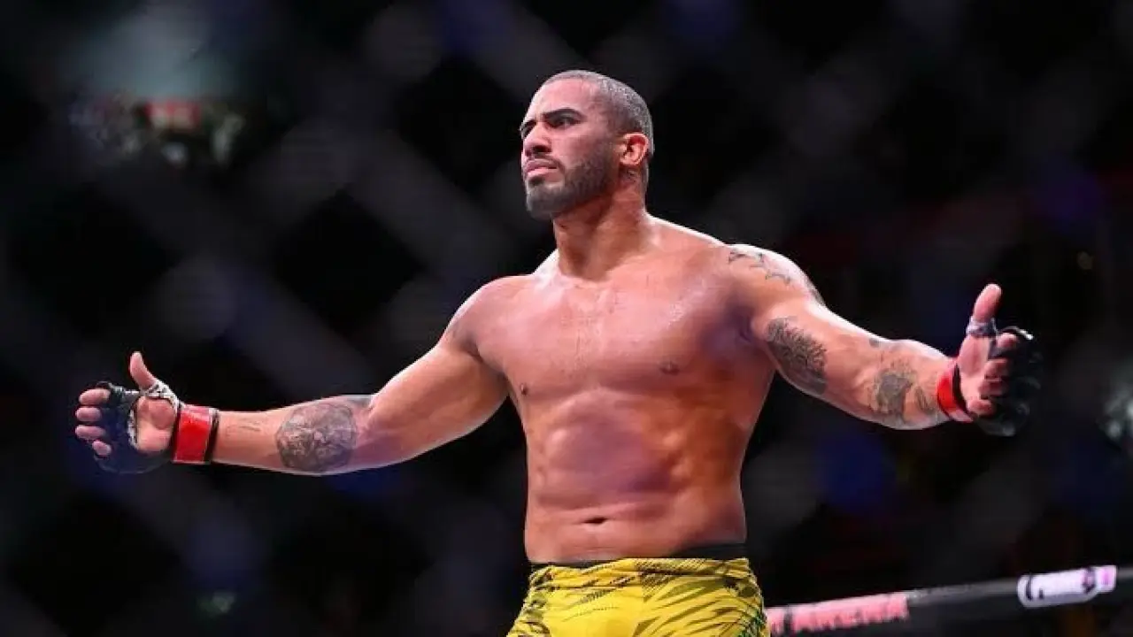 Lutador shirtless de braços abertos, com luvas vermelhas e calção amarelo, comemora dentro do octógono com a grade ao fundo