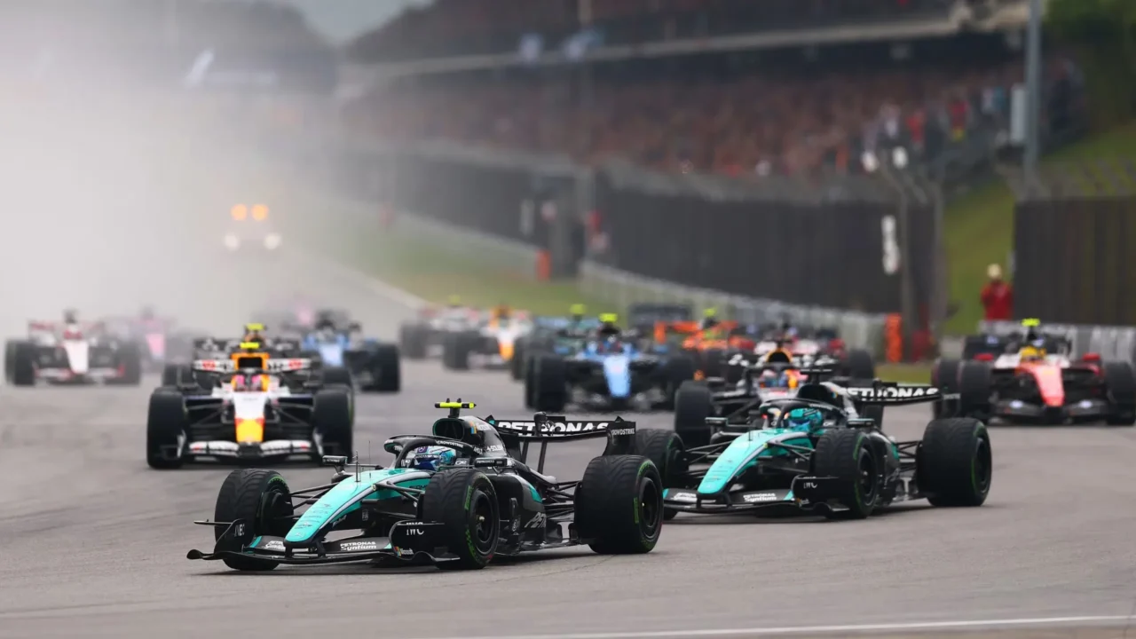 Monoposto de Fórmula 1 em turquesa lidera a largada de corrida em pista, com demais competidores atrás e fumaça dos pneus visível ao fundo, enquanto público ocupa arquibancadas do circuito