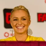 Mulher loira de brincos longos e blusa amarela sorri em painel diante de um fundo com logotipos vermelhos da FAN Expo