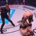 Lutador de camiseta preta e bermuda, de tatuagem no braço, aplica golpes por cima de outro lutador caído no chão do octógono, enquanto o árbitro observa de perto
