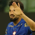 Jogador de camisa azul com escudo e patrocínios acena com a mão aberta em campo, com o rosto parcialmente encoberto pelo gesto