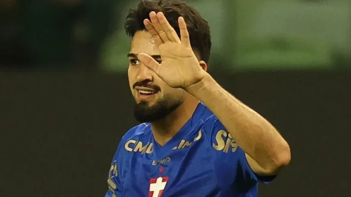Jogador de camisa azul com escudo e patrocínios acena com a mão aberta em campo, com o rosto parcialmente encoberto pelo gesto