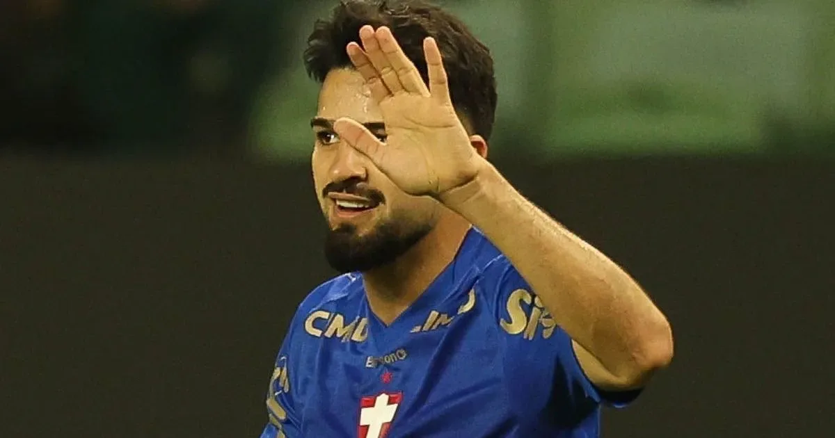 Jogador de camisa azul com escudo e patrocínios acena com a mão aberta em campo, com o rosto parcialmente encoberto pelo gesto