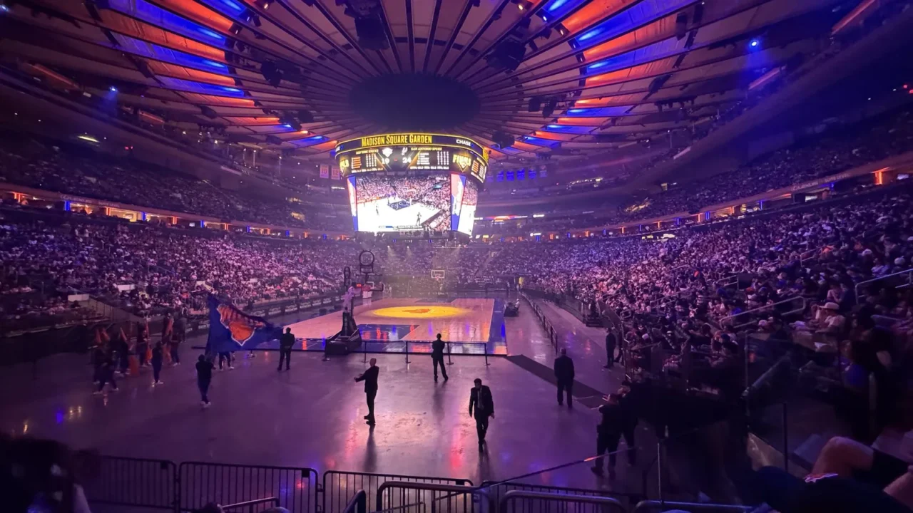 Madison Square Garden lotado, com luzes azuis e laranja no teto e a quadra iluminada, durante evento dos Knicks nas finais de 2026