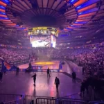 Madison Square Garden lotado, com luzes azuis e laranja no teto e a quadra iluminada, durante evento dos Knicks nas finais de 2026