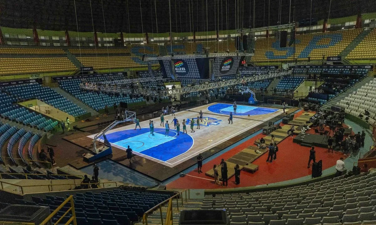 Ginásio do Ibirapuera preparado para partida de basquete com quadras azuis montadas no piso, equipes aquecendo e equipes de produção organizando o espaço para o jogo