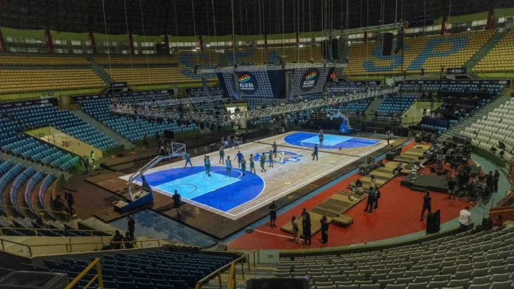 Ginásio do Ibirapuera preparado para partida de basquete com quadras azuis montadas no piso, equipes aquecendo e equipes de produção organizando o espaço para o jogo