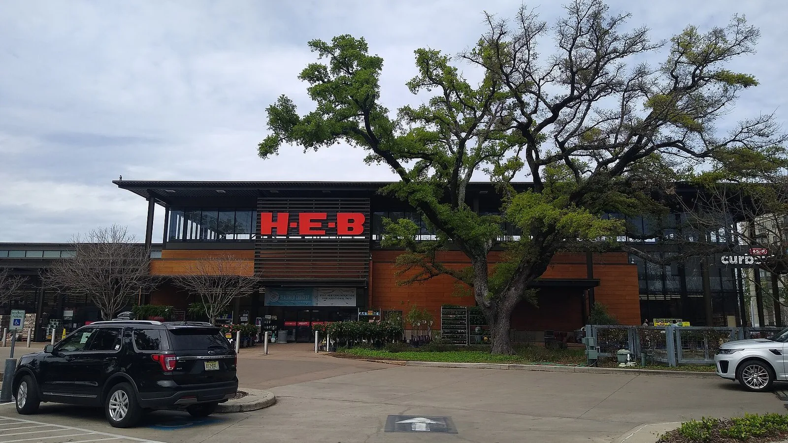 Entrada de uma loja H-E-B com letreiro vermelho, uma árvore grande e um carro preto no estacionamento