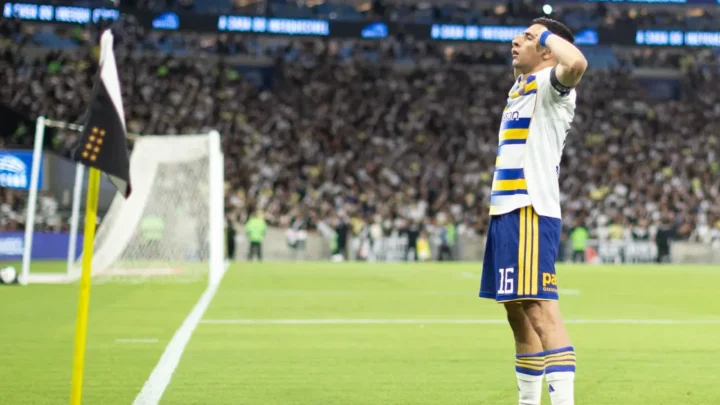 Jogador de camisa azul e amarela com listras, braço erguido à cabeça, comemorando perto da bandeira de escanteio em estádio lotado