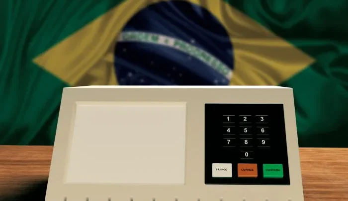Urna eletrônica diante da bandeira do Brasil. Cerca de 265 mil eleitores brasileiros registrados nos Estados Unidos votam neste domingo, 4 de outubro, no primeiro turno das Eleições 2026.