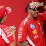 Piloto de camiseta vermelha da Ferrari, com óculos escuros e a mão na testa, conversa com uma integrante da equipe no paddock de um Grande Prêmio