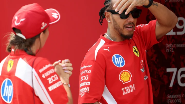 Piloto de camiseta vermelha da Ferrari, com óculos escuros e a mão na testa, conversa com uma integrante da equipe no paddock de um Grande Prêmio