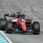 Carro vermelho de Fórmula 1, com pintura da Ferrari, acelera em pista de asfalto ao lado de zebras azuis e brancas na curva