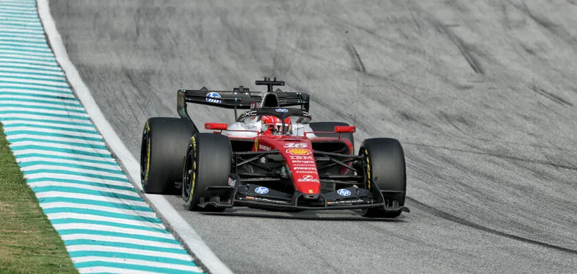 Carro vermelho de Fórmula 1, com pintura da Ferrari, acelera em pista de asfalto ao lado de zebras azuis e brancas na curva