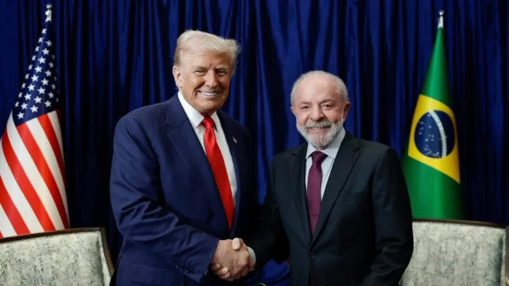 Dois homens apertam as mãos diante de bandeiras dos EUA e do Brasil e de cortina azul