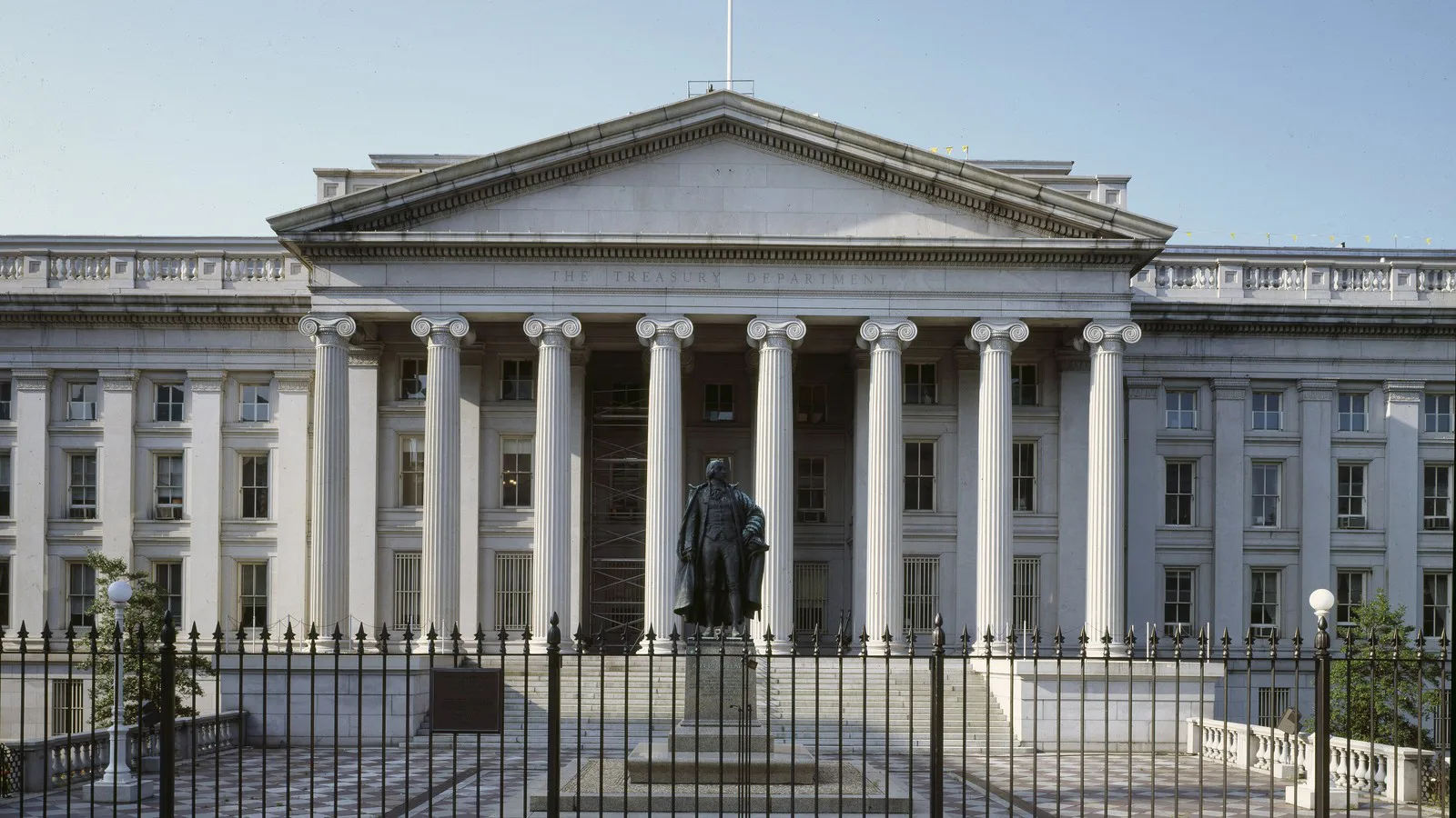Fachada do prédio do Departamento do Tesouro dos EUA, em Washington, com colunas e uma estátua à frente, atrás de uma grade