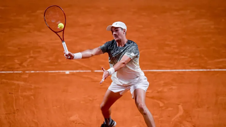 Tenista de boné branco e camiseta cinza devolve bola de backhand em quadra de saibro laranja, com raquete vermelha em ação