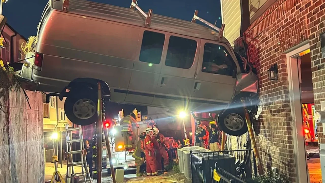 Van prateada suspensa entre um muro de contenção e a parede de tijolos de um sobrado, escorada por hastes, enquanto bombeiros trabalham embaixo, à noite, em Acworth, na Geórgia
