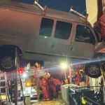 Van prateada suspensa entre um muro de contenção e a parede de tijolos de um sobrado, escorada por hastes, enquanto bombeiros trabalham embaixo, à noite, em Acworth, na Geórgia