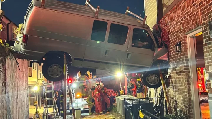 Van prateada suspensa entre um muro de contenção e a parede de tijolos de um sobrado, escorada por hastes, enquanto bombeiros trabalham embaixo, à noite, em Acworth, na Geórgia