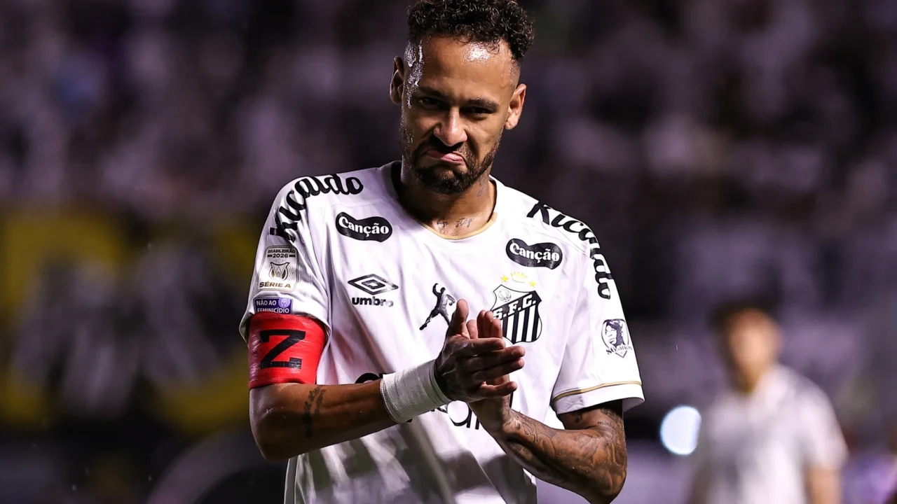 Jogador de camisa branca do Santos, com faixa de capitão vermelha, bate palmas com expressão de contrariedade durante partida em estádio à noite