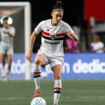 Jogadora de futebol feminino controla a bola durante partida do Brasileirão, usando uniforme branco com faixa vermelha e preta e patrocinador Superbet